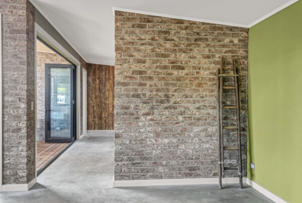 A room with brick walls, one painted green, includes a wooden ladder standing against the wall. Glass doors and windows are visible in the background, leading to another area.