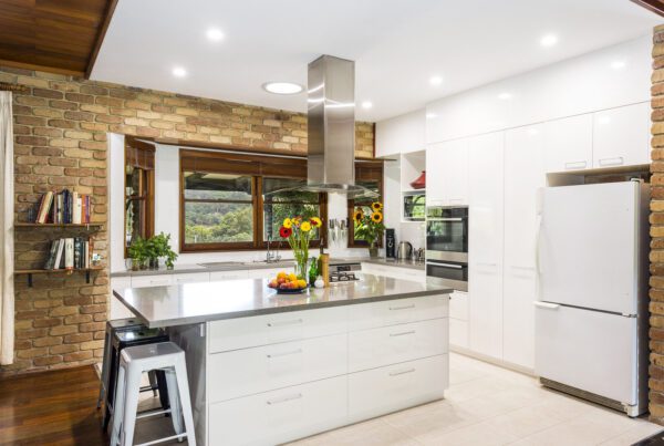 A modern kitchen with brick walls, white cabinets, stainless steel appliances, an island counter with fruit and flowers, and large windows overlooking greenery.