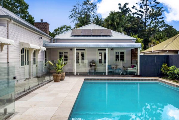 A backyard view of an Architect Mullumbimby-designed house showcases solar panels on the roof, a swimming pool in the foreground, and potted plants on a tiled patio. The property is enveloped by trees and lush greenery.